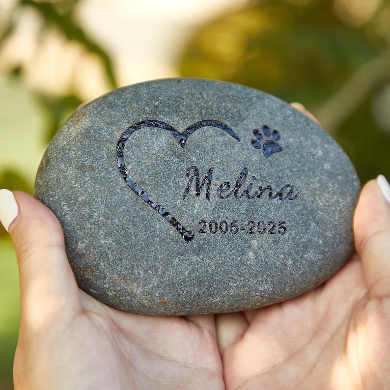 May include: A gray memorial stone with a heart and paw print design. The name "Melina" and the years "2005-2025" are engraved on the stone. The stone is held in two hands.