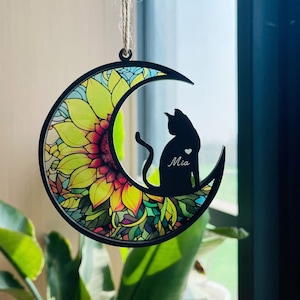May include: A black silhouette of a cat sitting on a crescent moon with a sunflower design. The cat's name, "Mia", is engraved on the moon.