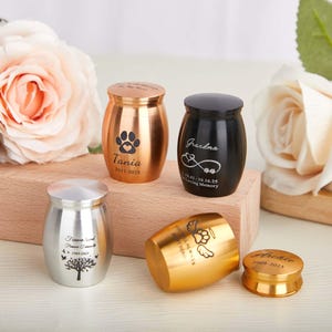 Op de afbeelding: Een verzameling kleine, metalen dierenurnen en gedenkwaardigheden in goud, zilver en zwart. De urnen hebben gegraveerde pootafdrukken, namen, data en liefdevolle boodschappen. De urnen worden tentoongesteld op een houten blok met zachtroze rozen op de achtergrond.