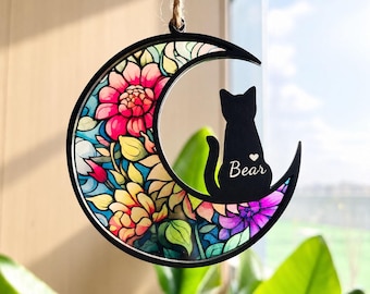 May include: A crescent moon-shaped hanging ornament with a black frame. The interior features a colorful floral pattern in shades of pink, yellow, and blue. A black silhouette of a cat sits on the moon with the name Bear and a heart.