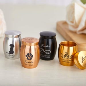 Puede incluir: Una colección de pequeñas urnas metálicas en plata, cobre, negro y oro. Las urnas presentan diseños grabados, incluyendo una silueta de perro, huellas de patas y texto. Una urna dorada tiene la tapa quitada.