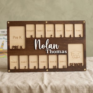 May include: A wooden school years photo display board with spaces for photos from Pre-K to Senior year. The board is stained dark brown with the name "Nolan Thomas" in white lettering. The word "SENIOR" is also visible.