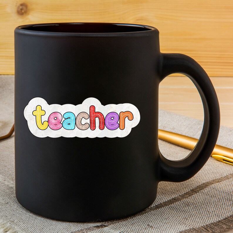 May include: Black coffee mug with a white sticker that says "teacher" in colourful letters.