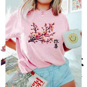 Op de afbeelding: Een roze T-shirt met een bloemenprint en het Japanse woord "Sakura" gedrukt op de voorkant. Het ontwerp toont een tak met kersenbloesems in roze en geel.