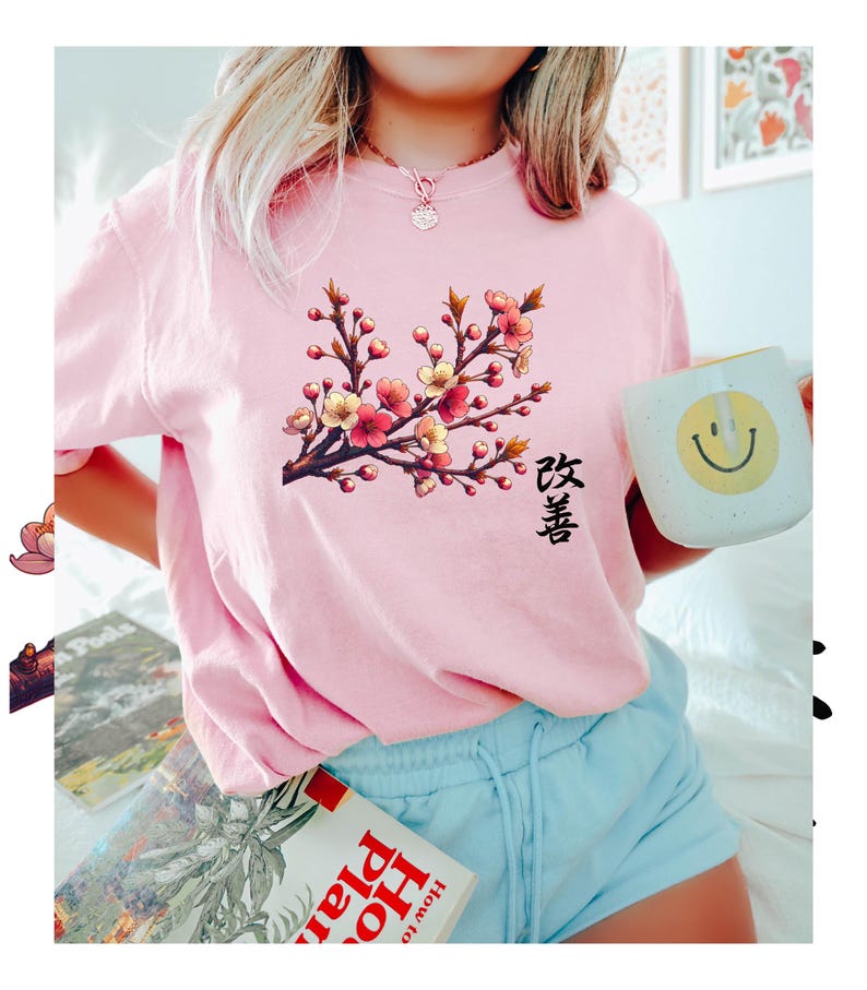 Op de afbeelding: Een roze T-shirt met een bloemenprint en het Japanse woord "Sakura" gedrukt op de voorkant. Het ontwerp toont een tak met kersenbloesems in roze en geel.
