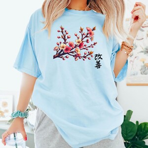 Op de afbeelding: Een lichtblauw t-shirt met een afbeelding van een roze en gele kersenbloesemtak met het Japanse woord "Sakura" gedrukt onder de tak.