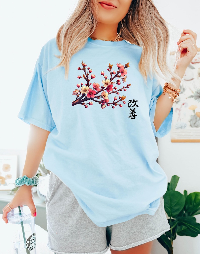 Op de afbeelding: Een lichtblauw t-shirt met een afbeelding van een roze en gele kersenbloesemtak met het Japanse woord "Sakura" gedrukt onder de tak.