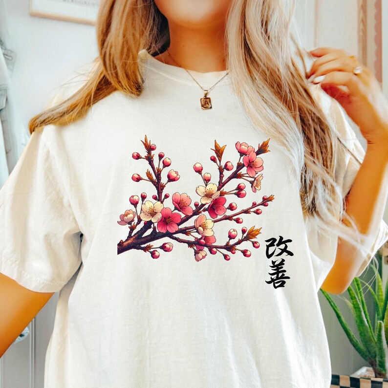 Op de afbeelding: Wit T-shirt met een roze en witte kersenbloesemtak ontwerp en het Japanse woord "Sakura" gedrukt in de rechter benedenhoek.
