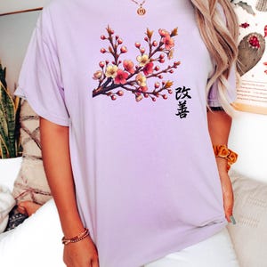 Op de afbeelding: Een lavendelkleurig T-shirt met een bloemenprint en het Japanse woord "桜" (Sakura) dat op de voorkant is gedrukt.