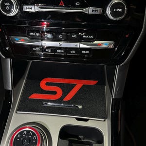 May include: A black car interior with a red ST logo on a black mat. The car has a gear shifter with a red ring around it and a cup holder.