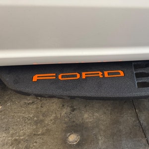 May include: A black step with a textured surface and a black metal grate. The word "FORD" is printed in orange on the step.