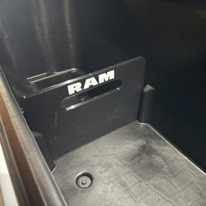 May include: Black plastic storage bin with a black metal divider. The divider has the word "RAM" in white letters. The bin is inside a vehicle.