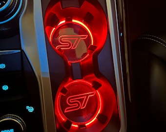 May include: Two red illuminated cup holders with the Ford ST logo in a car interior.