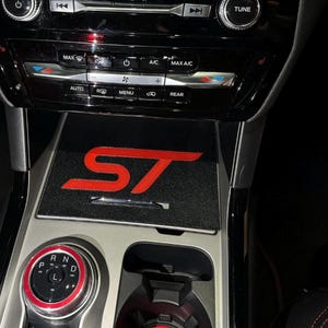 Puede incluir: Consola de coche negra con el logotipo "ST" rojo sobre una alfombrilla negra. La consola tiene botones para el control del clima, la radio y otras funciones. La palanca de cambios es roja y negra.