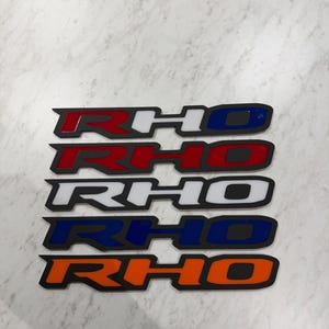 May include: Four RHO logo stickers in red, white, blue, and orange. Each sticker has a black border and a glossy finish. The stickers are arranged vertically on a light gray surface.