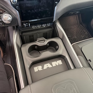May include: Interior view of a car's center console. The console is gray and black, featuring cup holders, a storage compartment, and a padded armrest with the RAM logo. A large infotainment screen is visible.