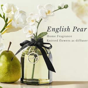 May include: A decorative home fragrance diffuser with the text "English Pear". The glass bottle contains a yellow liquid and is adorned with a black ribbon and a gold pendant. Knitted white flowers serve as the diffuser.