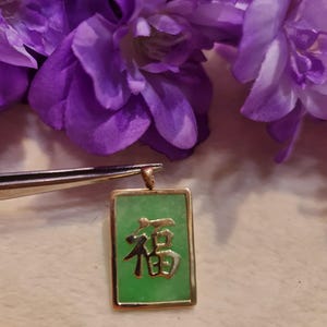 May include: A gold-colored rectangular pendant with a green jade center and a Chinese character. The pendant is held by tweezers, with purple flowers in the background. The jade pendant is approximately 1 inch long.