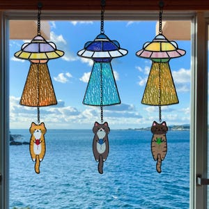 Op de afbeelding: Drie glas-in-lood UFO windgong met kattenhangers die voor een raam hangen met uitzicht op de oceaan. Elke UFO heeft een ander kleurenschema, waarbij de katten verschillende items vasthouden. De windgongs zijn opgehangen aan kettingen.