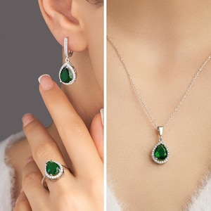 Puede incluir: Un conjunto de joyas de plata que incluye un anillo, un collar y pendientes. Cada pieza presenta una piedra preciosa verde esmeralda en forma de pera, rodeada por un halo de pequeñas piedras transparentes. Los pendientes son colgantes y el collar tiene una cadena fina.