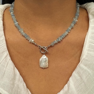 May include: A necklace featuring a strand of small, light blue, irregular gemstone chips. The necklace includes a silver-toned metal toggle clasp at the front, from which hangs a single, large, white, irregular-shaped baroque pearl pendant.