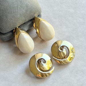 Puede incluir: Un par de pendientes de clip de tono dorado. Un par presenta una piedra ovalada blanca, el otro par es un diseño de espiral.