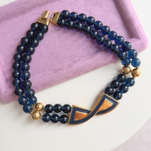 May include: A vintage necklace featuring two strands of dark blue beads. The necklace has a gold-tone clasp and a geometric pendant with a navy blue enamel finish and gold accents. The necklace is displayed on a lavender tray.