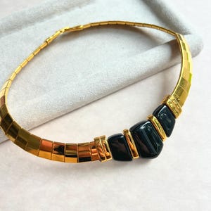 May include: A gold-toned necklace with a geometric design. The necklace features a series of rectangular links and is accented with black, rectangular stones. The necklace is displayed on a light gray surface.