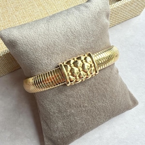 May include: A gold-colored bracelet with a textured, ribbed design. The center features a decorative, floral-like pattern. The bracelet is displayed on a gray, velvet-like pillow.