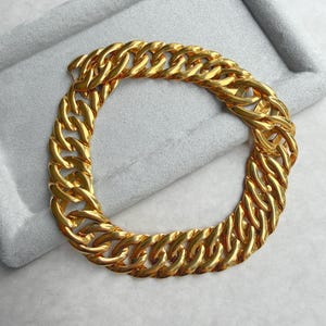 May include: A gold-colored chain bracelet with a thick, interlocking link design. The bracelet is displayed on a gray surface, showcasing its shiny, metallic finish. The chain links are closely connected, creating a bold and stylish accessory.