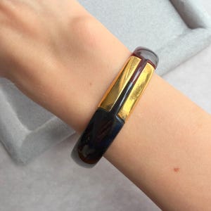 Puede incluir: Un brazalete azul oscuro y dorado. El brazalete tiene forma rectangular con un centro dorado y extremos azul oscuro. El brazalete se lleva en la muñeca.