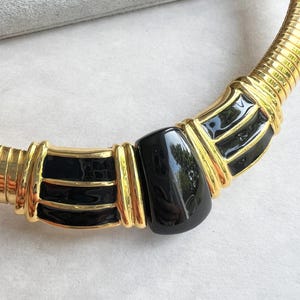 May include: A gold-toned collar necklace with a black enamel accent. The necklace features a flexible, ribbed design with a central black, rectangular element. The gold sections have a polished finish, creating a striking contrast with the black enamel.
