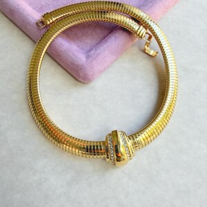 Könnte beinhalten: Eine goldfarbene Halskette mit geripptem Design. Die Halskette hat eine runde Form und einen Verschluss. Ein zentrales, rechteckiges Element ist mit kleinen, klaren Steinen verziert. Die Halskette wird auf einer hellvioletten Oberfläche präsentiert.