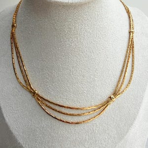 May include: A gold-colored necklace with three strands. The strands are connected by small, decorative clasps. The necklace is displayed on a white surface.