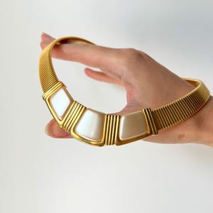 May include: A gold-toned choker necklace with a ribbed design. The necklace features five off-white, geometric-shaped segments framed in gold. The necklace is held by a hand against a white background.