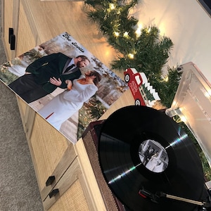 May include: A black vinyl record on a turntable with a wedding photo of a couple in front of a Christmas tree. The photo has the text "The Scotts" on it.