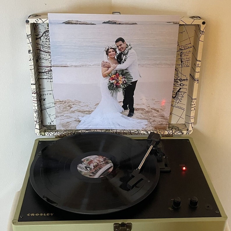 May include: A vintage-style record player with a black vinyl record on the turntable. The record player has a light green exterior with a map design and the word "CROSLEY" printed on the front. A photo of a couple on a beach is displayed on the record player's lid.