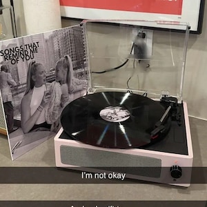 May include: A pink record player with a black vinyl record on the turntable. The record player has a clear plastic lid and a black knob on the side. The record player is sitting on a light brown surface. The record has the text "Songs that remind me of you" on the label.