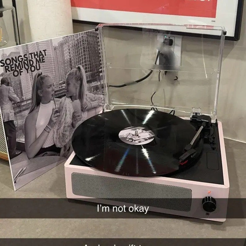 May include: A pink record player with a black vinyl record on the turntable. The record player has a clear plastic lid and a black knob on the side. The record player is sitting on a light brown surface. The record has the text "Songs that remind me of you" on the label.