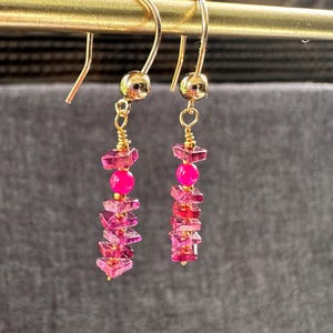 May include: A pair of dangle earrings with gold-colored hooks and accents. Each earring features a gold bead, a bright pink bead, and a series of small, faceted pink gemstones. The earrings hang from a gold bar against a gray background.