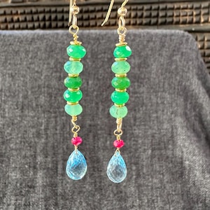 May include: Gold-toned dangle earrings featuring a series of green gemstone beads, small pink beads, and teardrop-shaped blue gemstones. The earrings are suspended from gold hooks and are displayed against a gray background.
