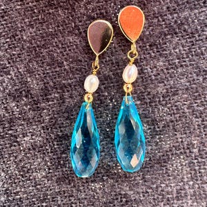May include: A pair of gold-toned earrings with a teardrop design. Each earring features a small, round top, a white pearl, and a large, faceted, blue-colored gemstone. The earrings are displayed against a dark, textured background.