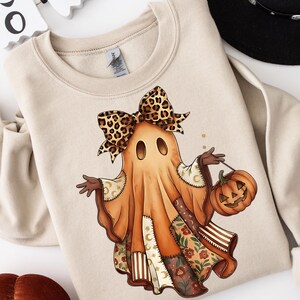 Könnte beinhalten: Beiges Sweatshirt mit einem Halloween-Geist-Charakter, Leopardenmuster-Schleife und einem geschnitzten Kürbis. Das Geisterkostüm hat Patchwork-Details mit Blumen- und Streifenmustern. Ein festliches und gemütliches Design.