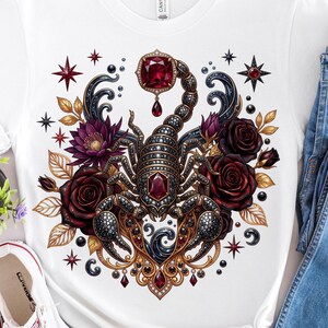 May include: White t-shirt featuring a detailed scorpion design with red gemstones, surrounded by dark red roses, gold leaves, and star accents. The scorpion is the focal point, with intricate detailing and a jewel-toned color palette.