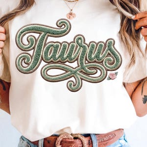 May include: Cream-colored t-shirt featuring the word "Taurus" in a green, textured, embroidered design. The word is in a stylized font with a decorative flourish. A small pink rose is next to the word.