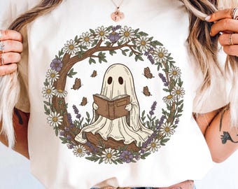 May include: Cream-colored t-shirt featuring a cartoon ghost reading a book, surrounded by a floral wreath of daisies, lavender, and butterflies. The ghost is draped in a sheet and has two black eye holes. The design is whimsical and nature-inspired.