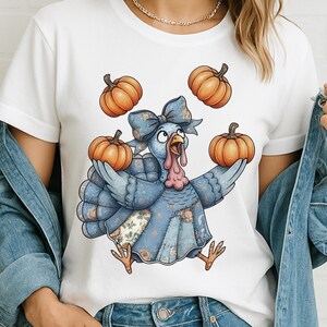 May include: White t-shirt with a cartoon turkey juggling three orange pumpkins. The turkey is blue with a floral pattern and a large blue bow. Ideal for Thanksgiving or autumn.
