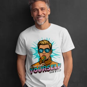 Può includere: T-shirt bianca con un'illustrazione in stile pop art di un uomo con occhiali da sole e un lecca-lecca. La grafica include il testo "The power to be YOURSELF every day" in un font colorato.
