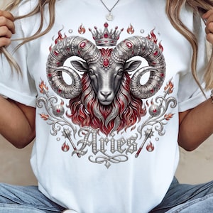 May include: White t-shirt featuring a detailed illustration of an Aries ram with ornate silver horns, a crown, and red accents. The word "Aries" is displayed in a decorative font below the ram.
