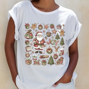 Könnte beinhalten: Weißes T-Shirt mit einem festlichen Weihnachtsdesign. Die Grafik zeigt den Weihnachtsmann, Geschenkboxen, Lebkuchenmänner, Weihnachtsbäume und das Wort "JOY". Das Design verwendet Rot-, Grün- und Brauntöne.